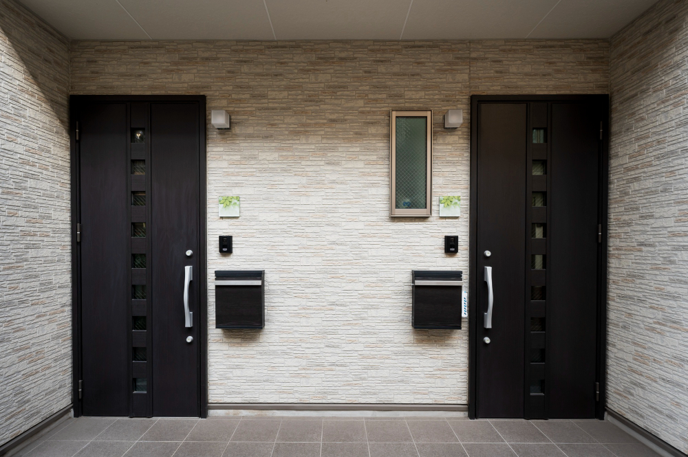 Modern apartment entrance with two dark solid core doors, mailboxes, intercom panels, and a small frosted window set in a light textured wall.