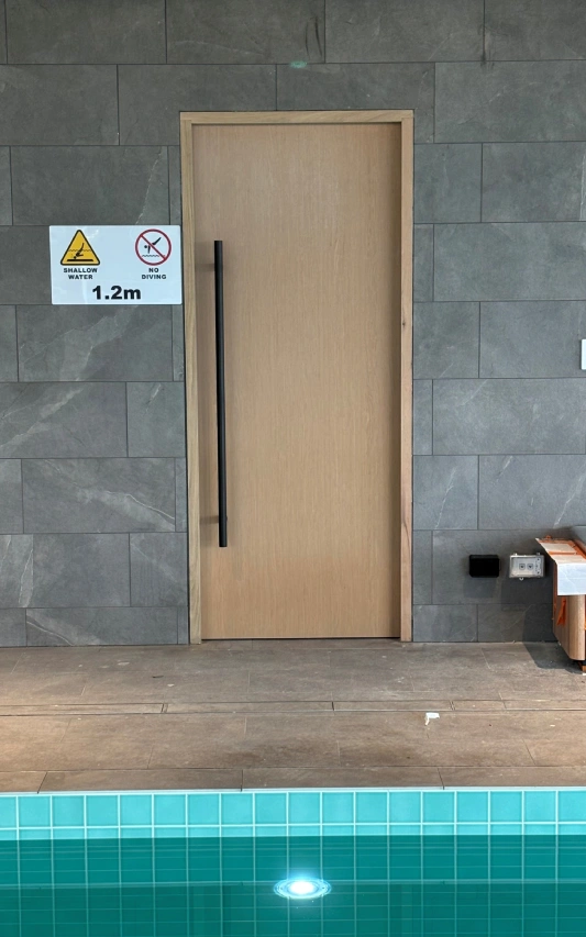Closed Tasmanian Oak door by Benefits Core Doors set in tiled wall near pool, warning sign and 1.2m marker.
