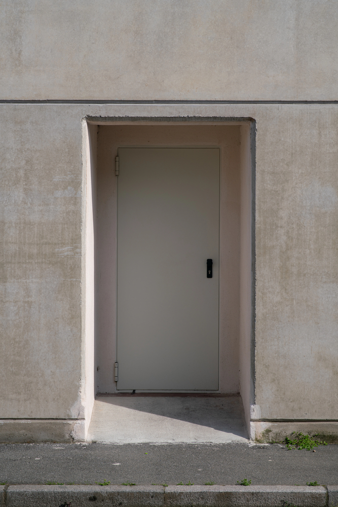 Closed gray fire door set in a recessed beige wall, with sidewalk and grass in front.