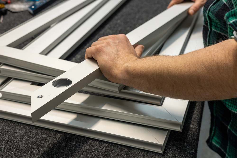 A person in a green plaid shirt assembles white steel door frames on a dark work surface.