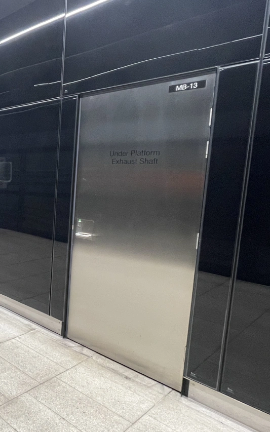 Metal door marked “Under Platform Exhaust Shaft” and code “MB-13” at tiled subway station platform.