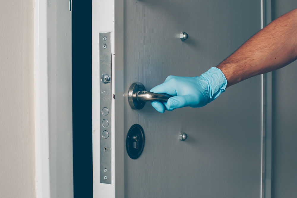 A hand in a blue nitrile glove opens a gray fire door, showing Brand compliance with New NSW Fire Regulations.