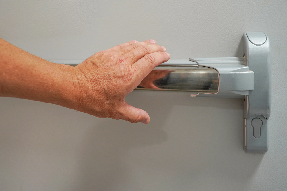 Hand pressing a metallic panic bar to perform a Common Fire Door Repairs inspection for maintenance purposes.